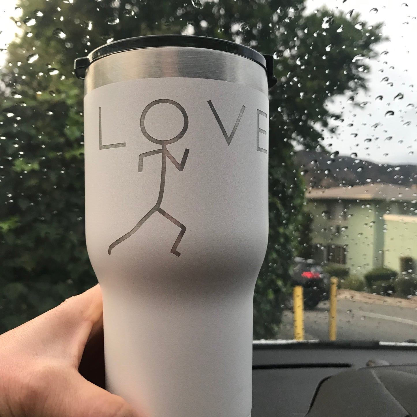 Coffee mug engraved with love design. the O in the word love is the head of a running stick figure. The background is a tree and some rain droplets on the windshield of the car.